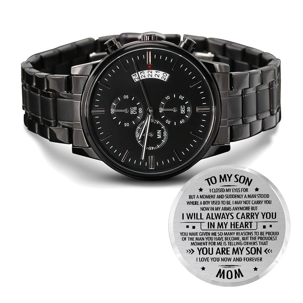 A black stainless steel chronograph watch with a copper dial, presented with an engraved message on a tag, indicating it is a gift from 'Mom' with options to personalize to 'Dad' or 'Mom and Dad'. The watch has a three-dial face, calendar function, and is water-resistant.